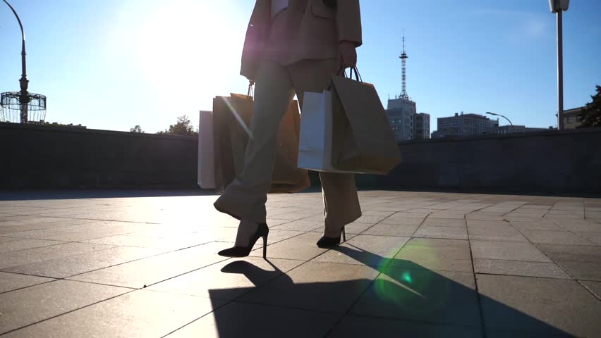 Fashionable woman carries paper packets going along urban street. Young female customer holds shopping bags walks at square after purchases. Concept of leisure at sales and discounts time. Slow motion