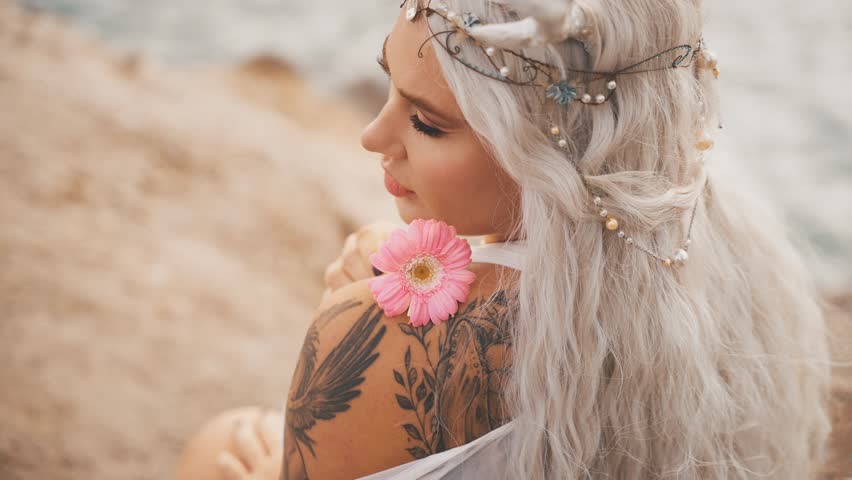 fantasy portrait young elf woman holding pink gerbera flower. close-up beauty face sexy eyes lips adult fairy girl, tiara diadem on head blonde ash hair. myth faun, pixie lady sits on riverbank sea.
