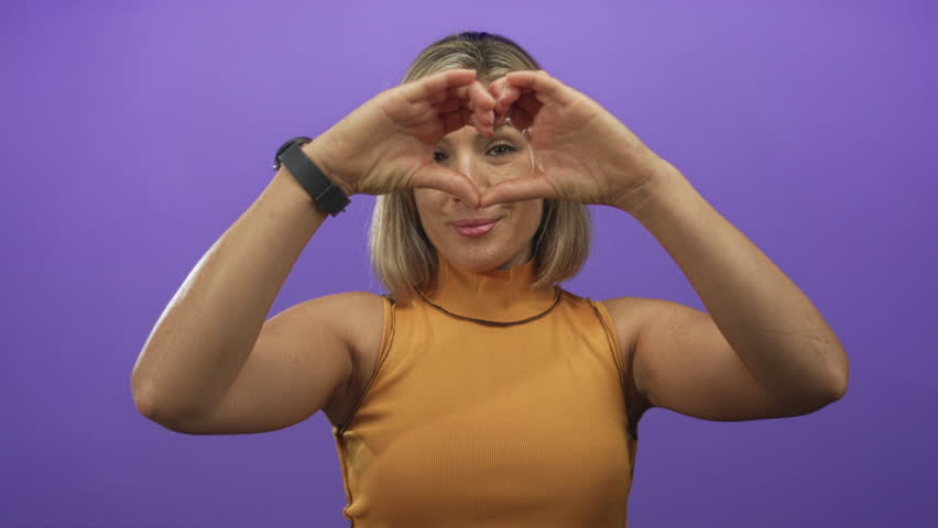 Woman makes heart shape with hands and holds them at chest level in a purple studio setting; love warmth.