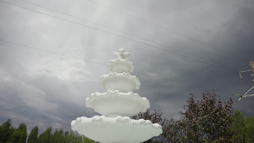 White decorative tiered fountain against a dramatic, cloudy sky with moody lighting and stormy weather vibes, creating a striking outdoor contrast.