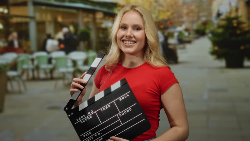 Woman holding clapperboard smiles on a bustling city street, blending creativity with urban life in a dynamic outdoor scene.