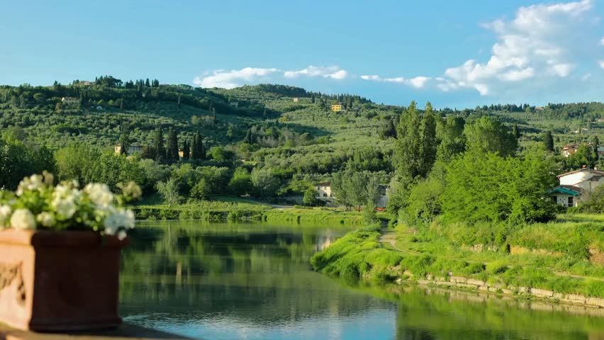 The serene Arno River winds through the vibrant, rolling hills of the Tuscan countryside in spring, dotted with cypress trees, blooming wildflowers, and rustic farmhouses under a soft blue sky.
