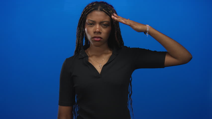 Woman standing saluting with right hand to forehead in blue studio, wearing black top with braids and cross necklace; determination respect.