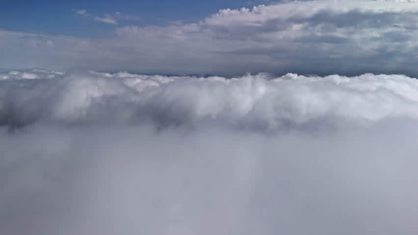 High altitude aerial view rises above a thick endless sea of dense white cumulus clouds with blue sky visible above