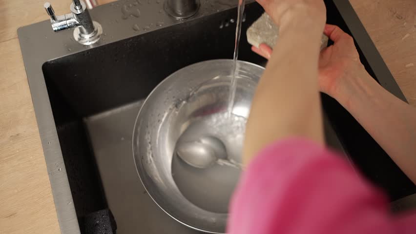 Female hands washing a stainless steel bowl and spoons with a sponge and soapy water in a modern black kitchen sink, focusing on the daily domestic chore of cleaning up after a meal