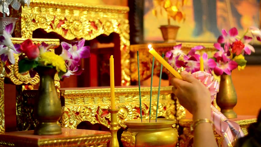 Person lighting a candle at a golden buddhist shrine with flowers and incense sticks