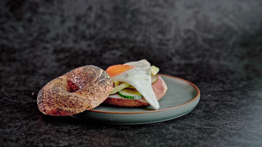 Close-up of male hand in glove sprinkling sesame on freshly made bagel. Teal plate on dark background. Food styling, final touch concept, delicious sandwich presentation.