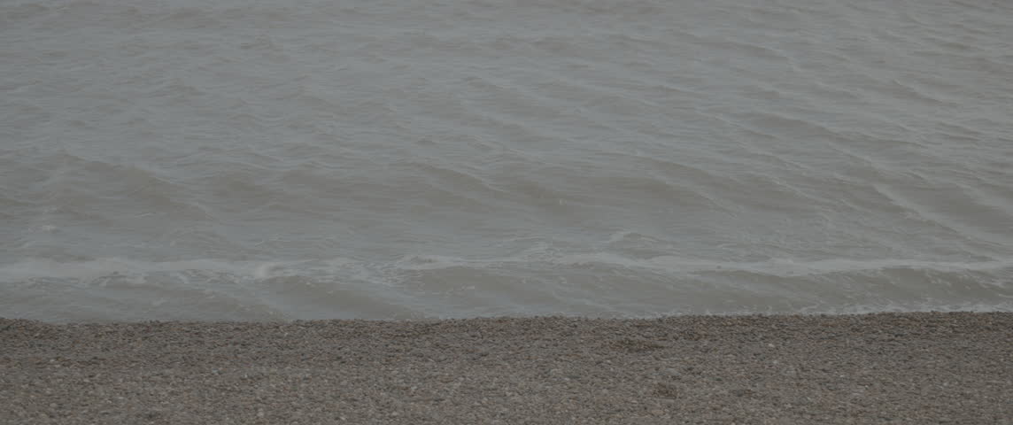 Slow-motion static mid shot of a single gentle wave crashing along the Suffolk shingle shoreline during the day