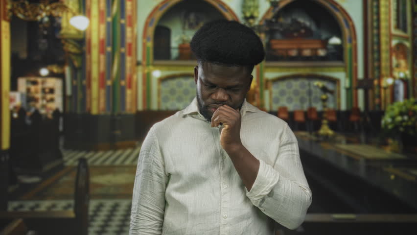 Young african american man hand on chin pensive pose inside ornate catholic church nave wearing light linen shirt and looking downward; quiet contemplation.