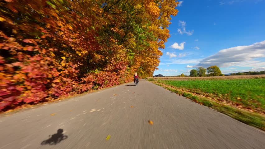 Cinematic FPV drone shot of woman riding motorcycle along country road under vibrant golden autumn trees. Warm light, cozy colors, and peaceful travel adventure mood.
