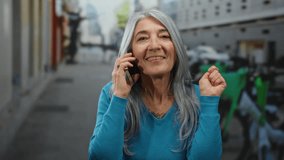 Senior woman with grey hair in blue sweater talks joyfully on a smartphone while standing in a lively urban street setting surrounded by bicycles and cityscape. - Powered by Shutterstock - Get 15% off with code: PIKWIZARD15