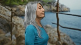 Senior woman with grey hair standing outdoors by the seaside at the beach, looking contemplatively beyond the rocky landscape with ocean in the background. - Powered by Shutterstock - Get 15% off with code: PIKWIZARD15