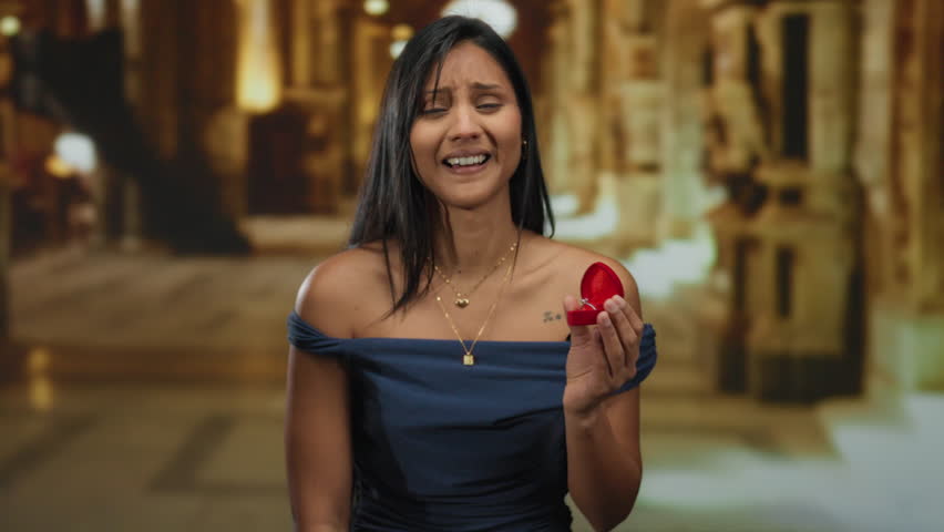 Woman holding red ring box in church setting indoors, expressing emotion with hispanic or latin heritage, wearing elegant attire and jewelry against a blurred background.