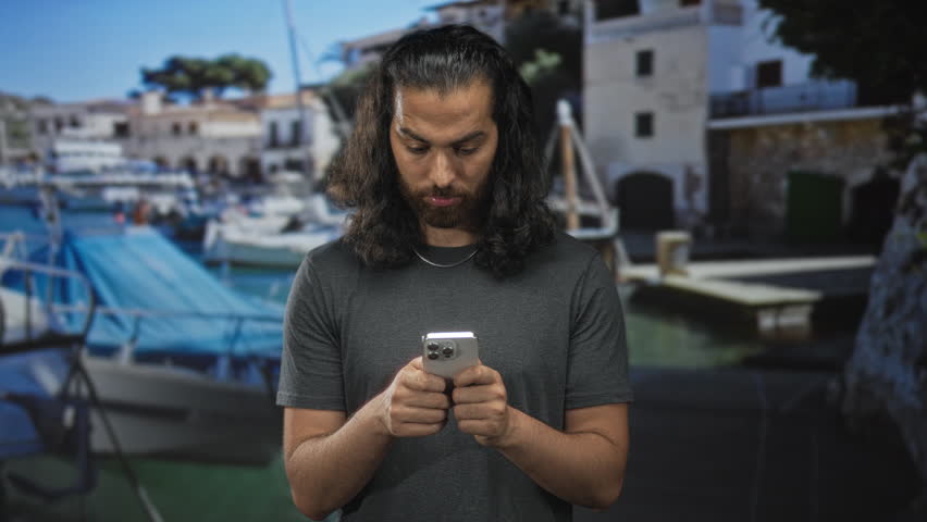 Man with long hair and beard looking down at smartphone, hands typing by a waterfront building with moored boats and wooden dock; quiet focus.