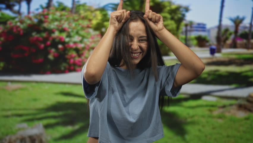 Woman smiling with fingers to head as horns, wearing cropped blue shirt in park building with green lawn and flowers; carefree joy.