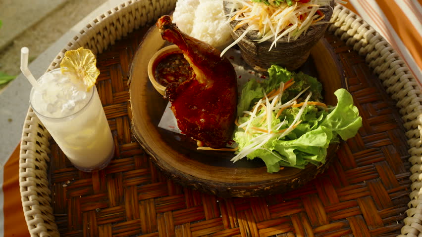 Overhead view of grilled chicken with sticky rice, papaya salad and lemonade on woven tray beside the sea, overhead, Thai Street Food Concept.