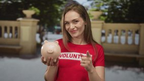 Woman in red volunteer shirt with outstretched finger points to blush ceramic piggy bank on busy urban street; doubt. - Powered by Shutterstock - Get 15% off with code: PIKWIZARD15