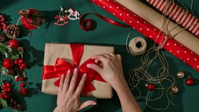 Female hands adjusting a red satin ribbon bow on a kraft paper wrapped Christmas present surrounded by festive decorations, and wrapping materials on a green background. - Powered by Shutterstock - Get 15% off with code: PIKWIZARD15