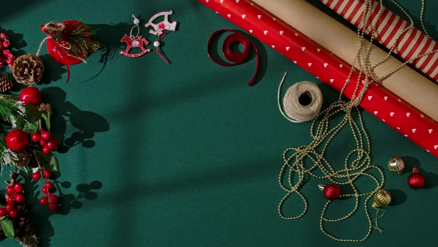 Top view of female hands holding a smartphone with a green screen on a dark green festive background. Holiday wrapping materials, ribbons, gold beads, red ornaments, and pine decorations surround the
