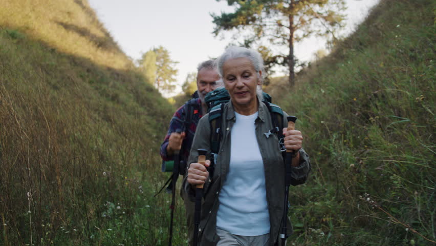 Senior woman with backpack walks through narrow grassy canyon and older man follows behind. Female grips trekking poles and steps firmly on winding dirt path