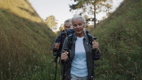 Senior woman with backpack walks through narrow grassy canyon and older man follows behind. Female grips trekking poles and steps firmly on winding dirt path - Powered by Shutterstock - Get 15% off with code: PIKWIZARD15