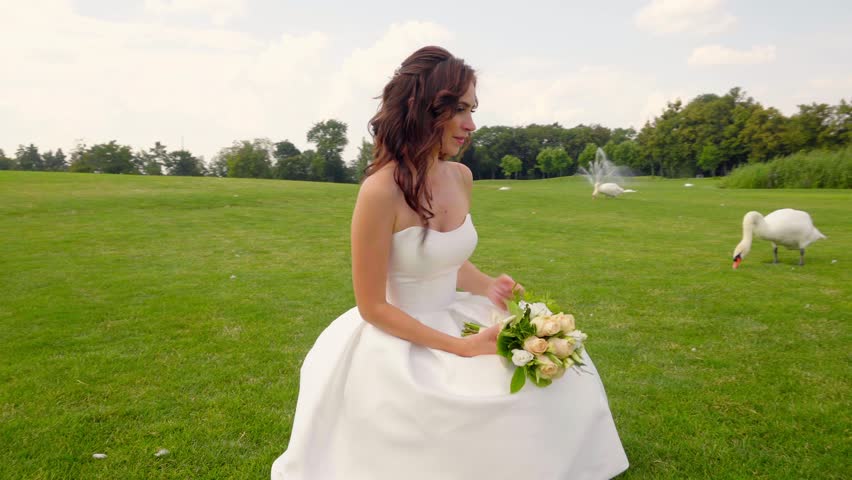 Young bride sitting on green grass, wearing a white wedding dress and holding a floral bouquet under sunny skies, representing love, joy, and outdoor celebration 4K UHD B-Roll footage