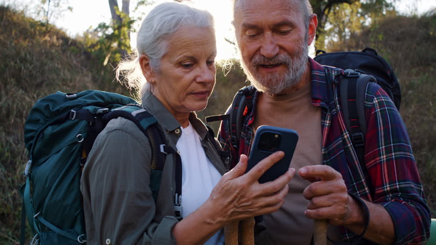Senior woman holds smartphone while mature man pointing ahead. Grey haired woman opens map app and taps screen setting path waypoint before walk starts