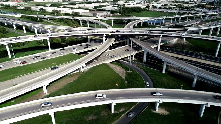 Dynamic aerial perspective showcasing a complex highway interchange filled with moving vehicles surrounded by greenery and urban structures.