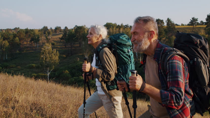 Positive elderly friends tourists go uphill in autumn valley slow motion. Mature woman shows interesting sight forward husband during Nordic walking in park
