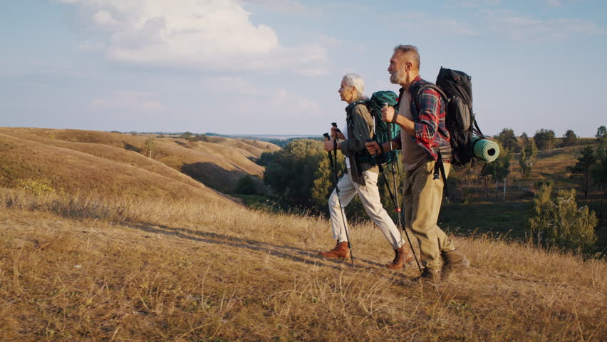 Inspiring elderly couple of hikers goes uphill in autumn valley slow motion. Athletic mature woman and man practice Nordic walking in nature park on sunny day
