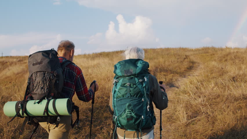 Trained old hikers with rucksacks go uphill in autumn valley rear view. Healthy elderly family of woman and man exercises in Nordic walking in sunny park