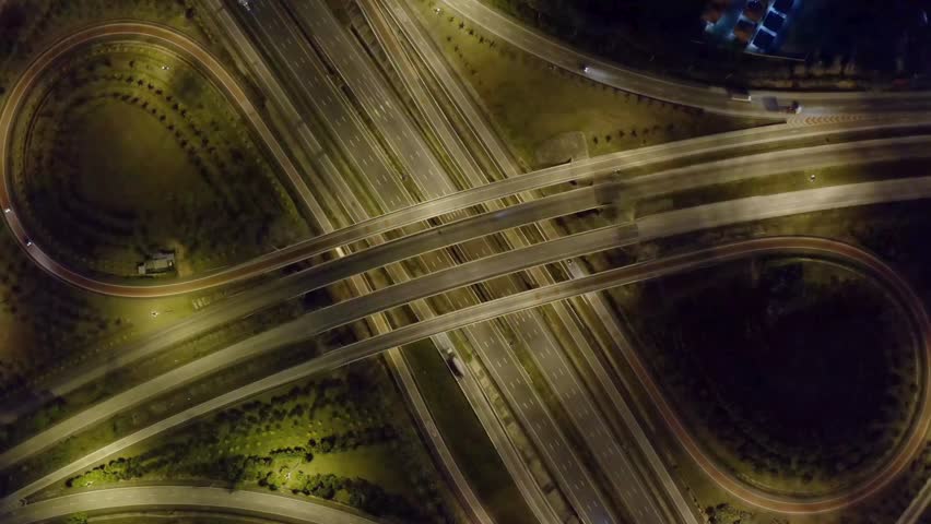 Nighttime aerial perspective reveals complex highway interchange with flowing traffic, surrounded by greenery and urban lighting enhancing the dynamic landscape.