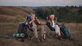 Senior couple sits on camping chairs on hill slope enjoying rest. Old man passes compact binoculars to woman explaining focus wheel during pause at Nordic walk - Powered by Shutterstock - Get 15% off with code: PIKWIZARD15