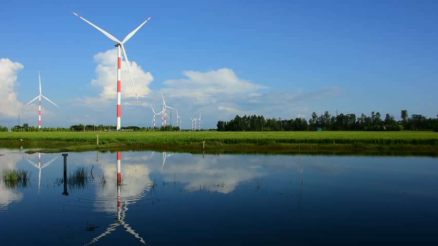 Wind turbines rotate beside a calm waterbody in coastal Bangladesh under a blue sky. A scenic video highlighting renewable energy and the harmony between nature and technology.