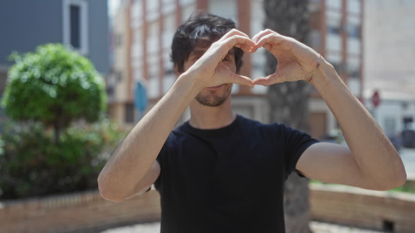 Man smiling and making heart shape with hands on street in black tshirt by green bush under bright sunlight; affection.