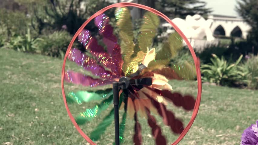 A colorful, handmade flower swirling and moving in the wind, alongside flowers in a green field. A tribute to respect and affection. Outdoors, with plenty of natural light.