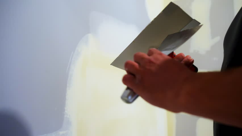 Construction worker spreading joint compound on the drywall wall using a metal taping knife. Wall smoothed and finished as part of a home renovation.