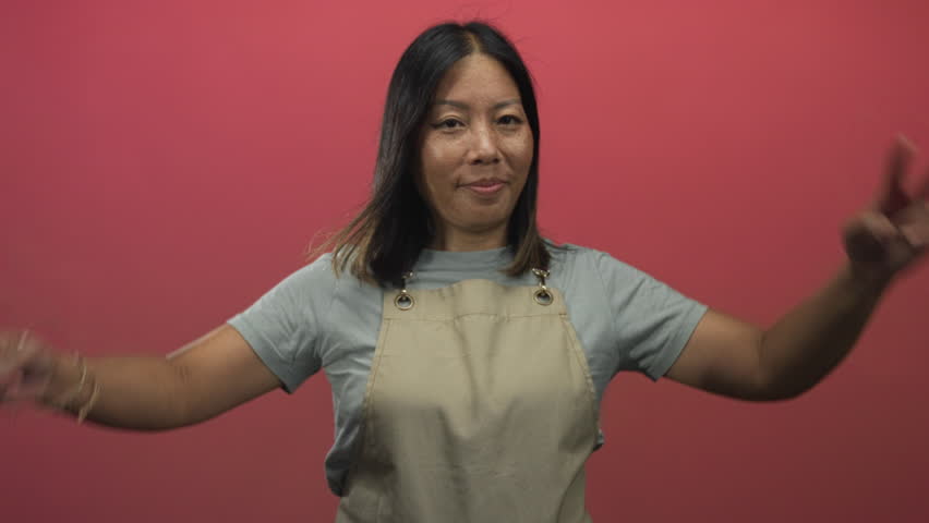 Woman showing double peace signs with both hands in pink studio wearing apron and grey t shirt while smiling broadly; playful friendly approachable.