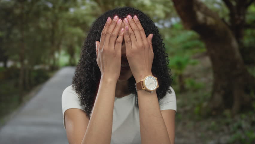 Young woman covers face with both hands on sunlit forest path lined with tall trees and green foliage; shyness.