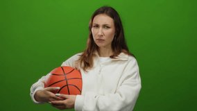 Woman holding basketball making quiet gesture against isolated green background in casual white outfit suggesting calm but focused attitude indoors. - Powered by Shutterstock - Get 15% off with code: PIKWIZARD15