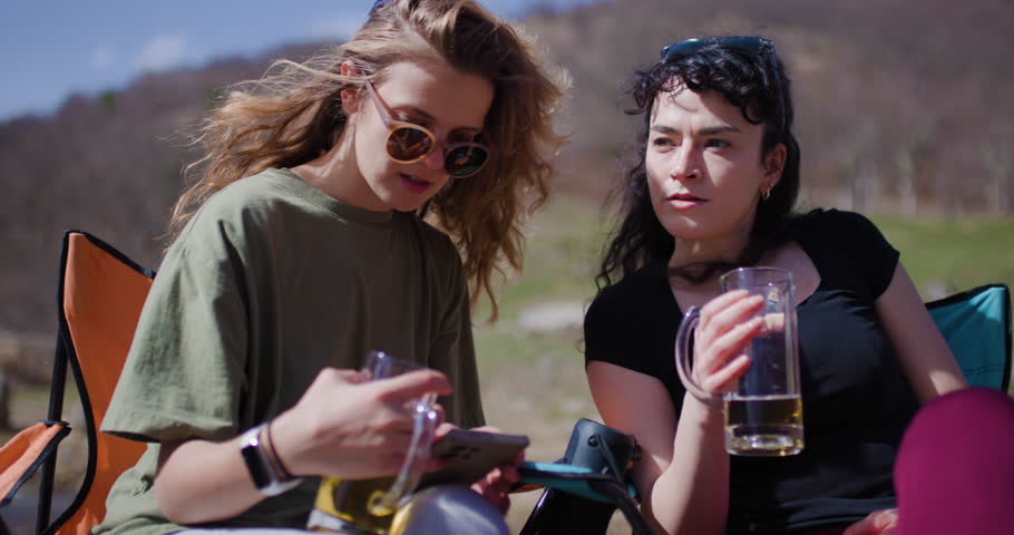 Two female friends relax in camping chairs by a stream, enjoying drinks and browsing a smartphone. They appear to be on a camping trip, enjoying the outdoors.