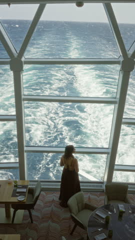 Hispanic young brunette woman standing by a large panoramic window looking at the ocean wake from a cruise ship inside building restaurant with table settings; serenity.