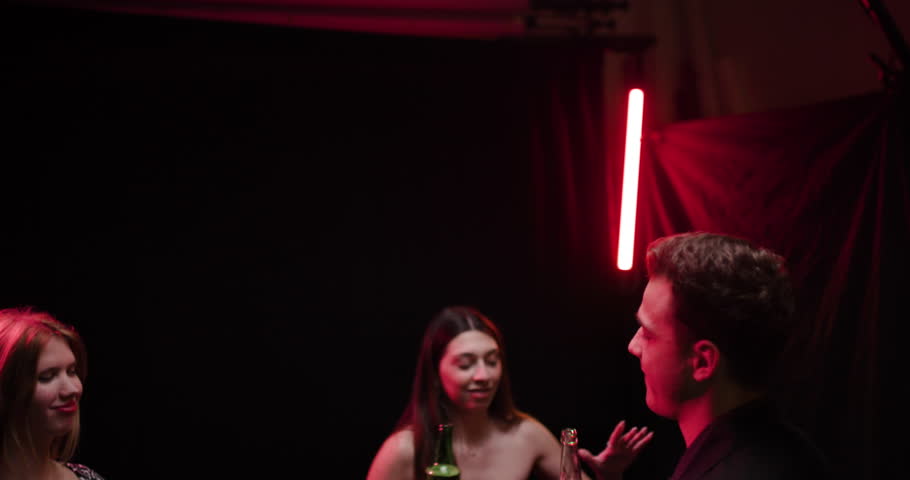 Friends enjoying drinks at a party, laughing, and having a good time. The atmosphere is lively and fun, with colorful neon lights and smoke.