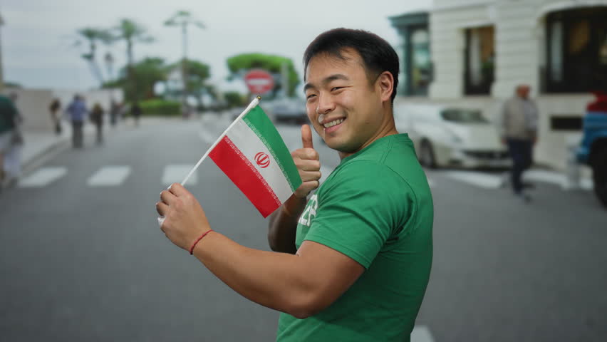 Young man holding iranian flag outdoors in urban street setting, showcasing cultural pride and diversity.