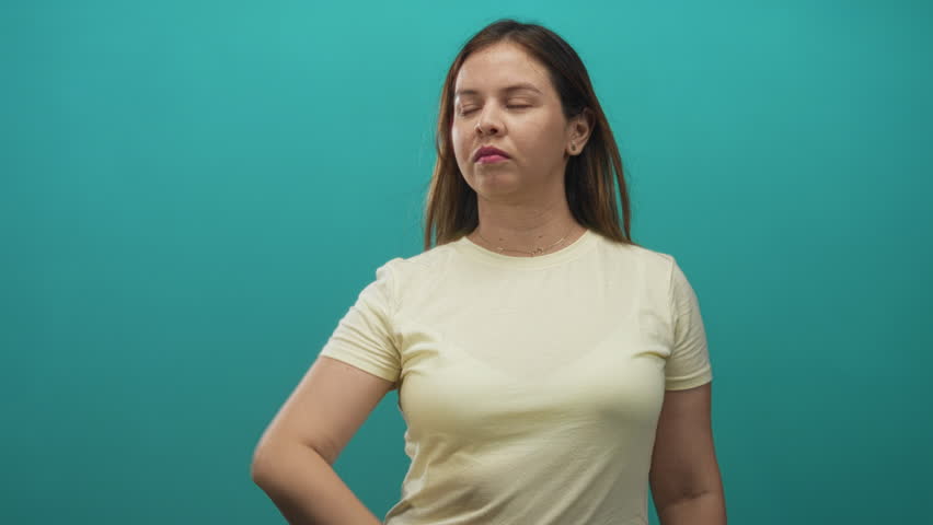 Woman holding palm up and looking to side in teal studio wearing pale yellow tshirt; mild skepticism.