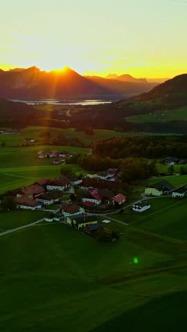 Vertical aerial parallax orbits idyllic Austrian village in green rolling hills during golden sunset over an Alpine mountain lake reflecting light