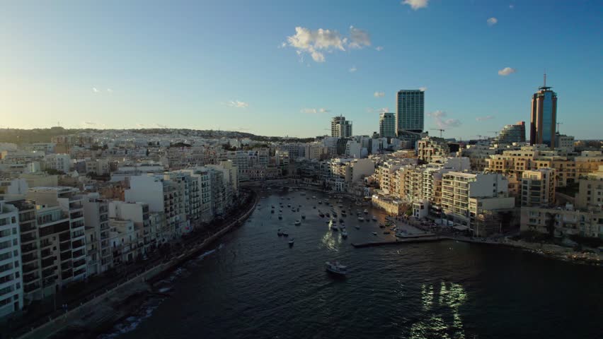 Aerial lateral shot of St. Julian’s Bay at sunset, showing the dense coastal cityscape, tall buildings, boats anchored in the bay, and golden reflections on the water.