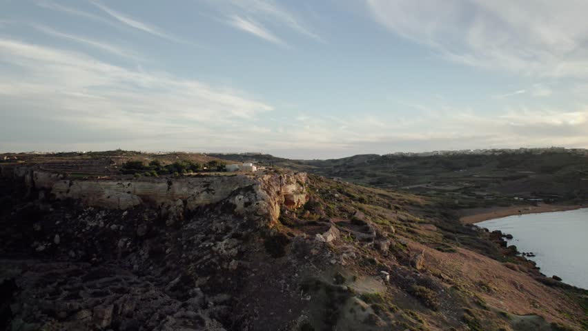 The drone flies straight toward the cliffs of Gozo, then rises to skim above them, following a winding road bathed in golden evening light.