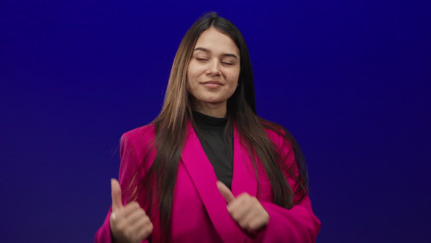 Young hispanic woman in pink points thumbs backward while smiling against a vibrant blue background, dressed in a fashionable jacket, highlighting happiness and style.