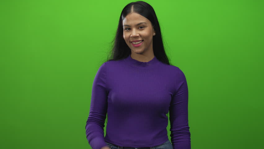 Young latin woman waving hand in studio green screen wearing purple top and denim jeans smiling at camera; friendliness.